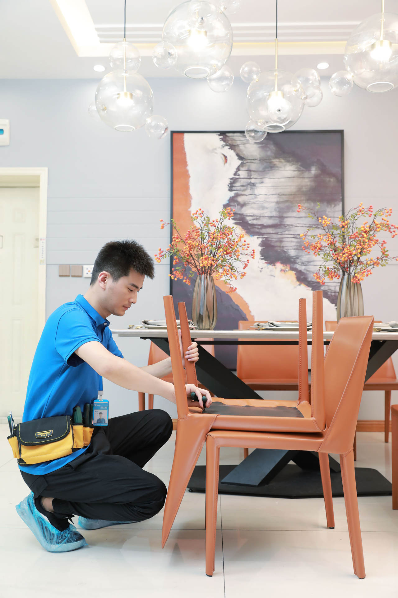 现代餐厅背景下，身穿蓝色制服的专业师傅正蹲地组装橙色皮质餐椅。他手持螺丝刀紧固椅腿螺丝，腰间佩戴印有WINHUNT字样的工具包，脚穿蓝色鞋套保护地面。背景可见白